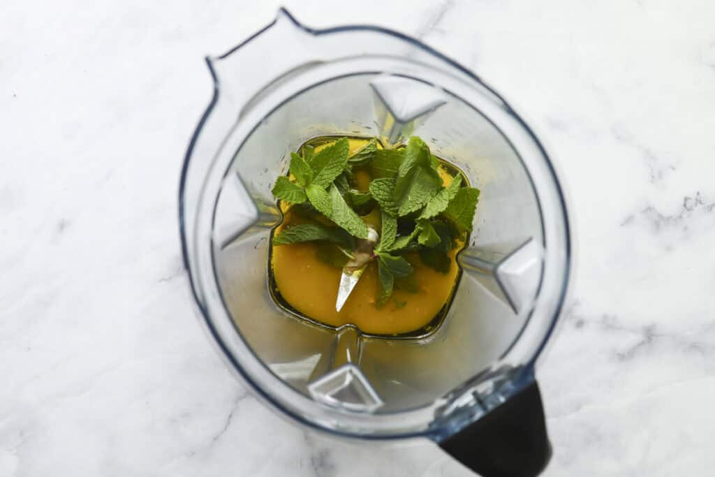 A top-down view of a blender with fresh mint leaves and an orange liquid inside, placed on a white marble surface.