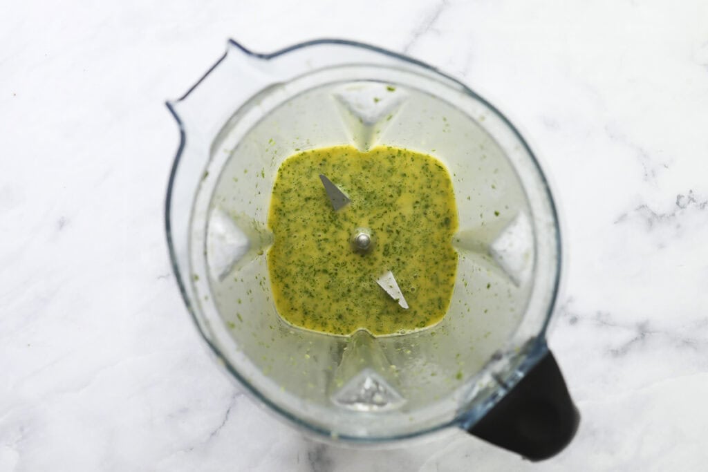 A top-down view of a blender containing a green and yellow blended mixture with visible herb flecks, sitting on a white marble surface.