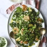 A white platter holds a shrimp & zucchini pasta salad with shrimp, kale, artichoke hearts, green peas, mint, and crumbled cheese. Two serving spoons rest on the plate. Nearby are a glass of white wine, dressing, and chopped herbs.