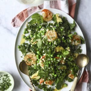 A white platter holds a shrimp & zucchini pasta salad with shrimp, kale, artichoke hearts, green peas, mint, and crumbled cheese. Two serving spoons rest on the plate. Nearby are a glass of white wine, dressing, and chopped herbs.