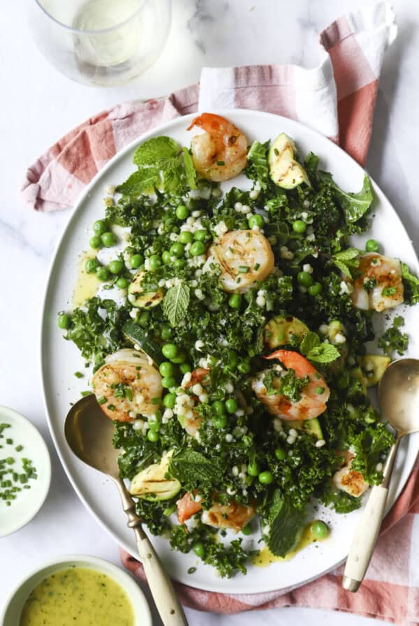 Shrimp & Zucchini Pasta Salad sits on a white platter with kale, green peas, grilled shrimp, zucchini, and fresh herbs, drizzled with dressing. Two serving spoons rest on the platter. A red and white cloth and bowls of dressing are nearby.