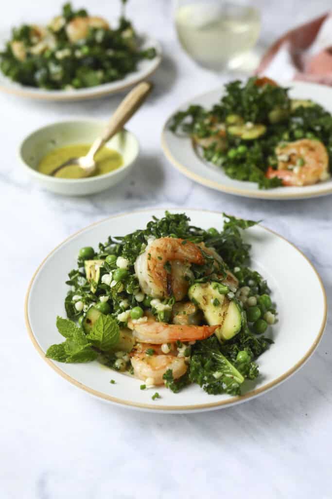 A plate of shrimp & zucchini pasta salad with shrimp, kale, couscous, peas, cucumber, and fresh mint, served with a small bowl of dressing. More salad plates are in the background on a white surface.