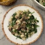 A plate of artichoke pasta salad with white beans, spinach, and fresh herbs sits on a marble surface next to a bowl of lemons and another filled with salad. The pasta is beautifully garnished with vibrant herbs.