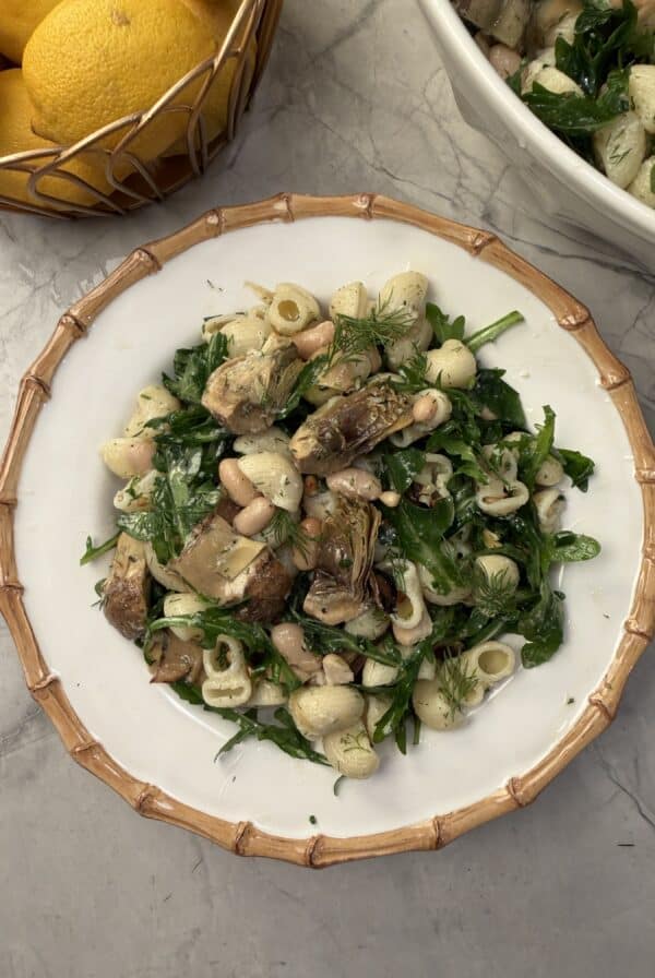 A plate of artichoke pasta salad with mushrooms, white beans, spinach, and fresh herbs is served on a white dish with a bamboo-patterned rim. A bowl of lemons and another bowl of salad rest nearby on a marble surface.