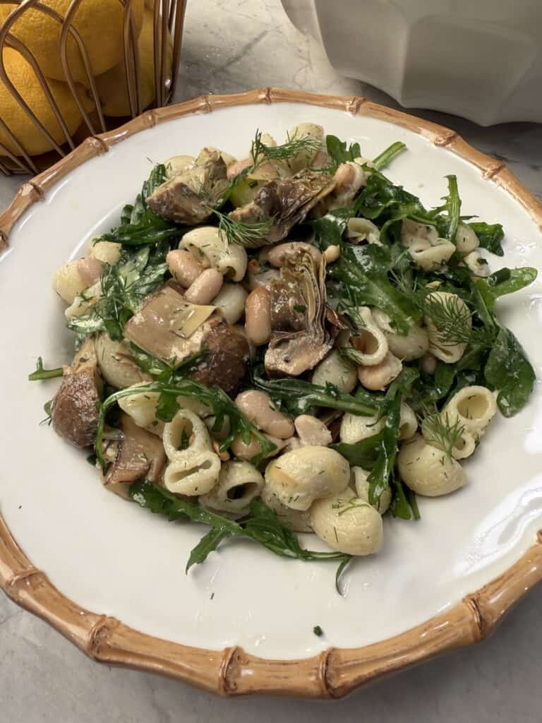 A plate of artichoke pasta salad with white beans, arugula, fresh dill, and a creamy dressing is served on a white plate with a tan rim. Lemons in a basket can be seen in the background.