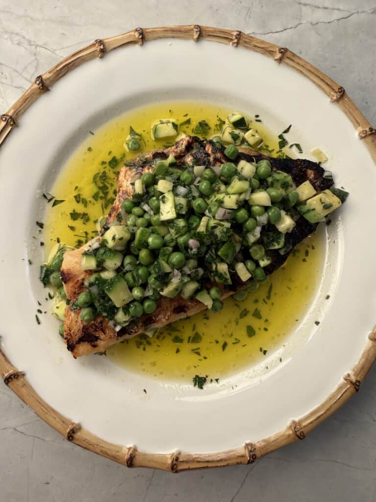 A Crispy Grilled Fish fillet topped with a green pea and diced cucumber salad, garnished with herbs and served in a pool of yellow oil or sauce on a white plate with a bamboo-like rim.