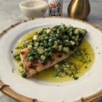 A crispy grilled fish fillet topped with diced cucumber, peas, and herbs sits in a pool of olive oil on a white plate. In the background are a gold oil dispenser, a small bottle, and a marble bowl.