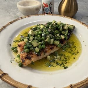 A crispy grilled fish fillet topped with diced cucumber, peas, and herbs sits in a pool of olive oil on a white plate. In the background are a gold oil dispenser, a small bottle, and a marble bowl.