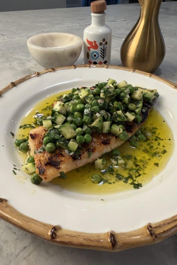 A crispy grilled fish fillet topped with diced cucumber, peas, and herbs sits in a pool of olive oil on a white plate. In the background are a gold oil dispenser, a small bottle, and a marble bowl.