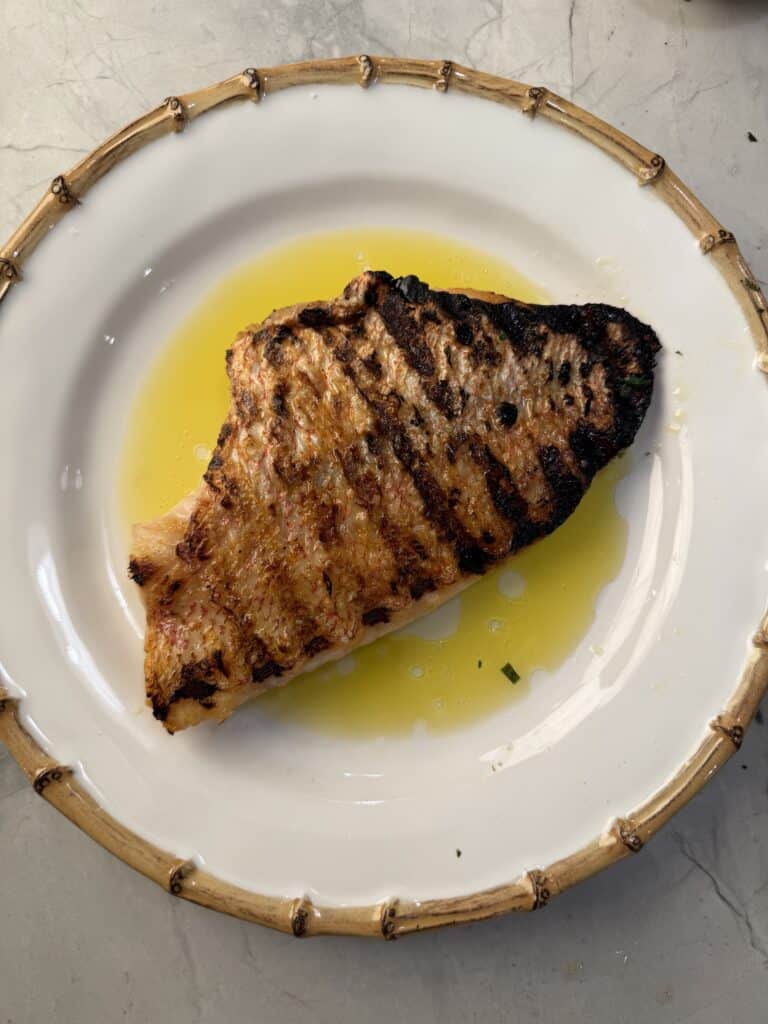 A crispy grilled fish fillet with char marks sits on a white plate with a bamboo-style rim, surrounded by a pool of golden olive oil.
