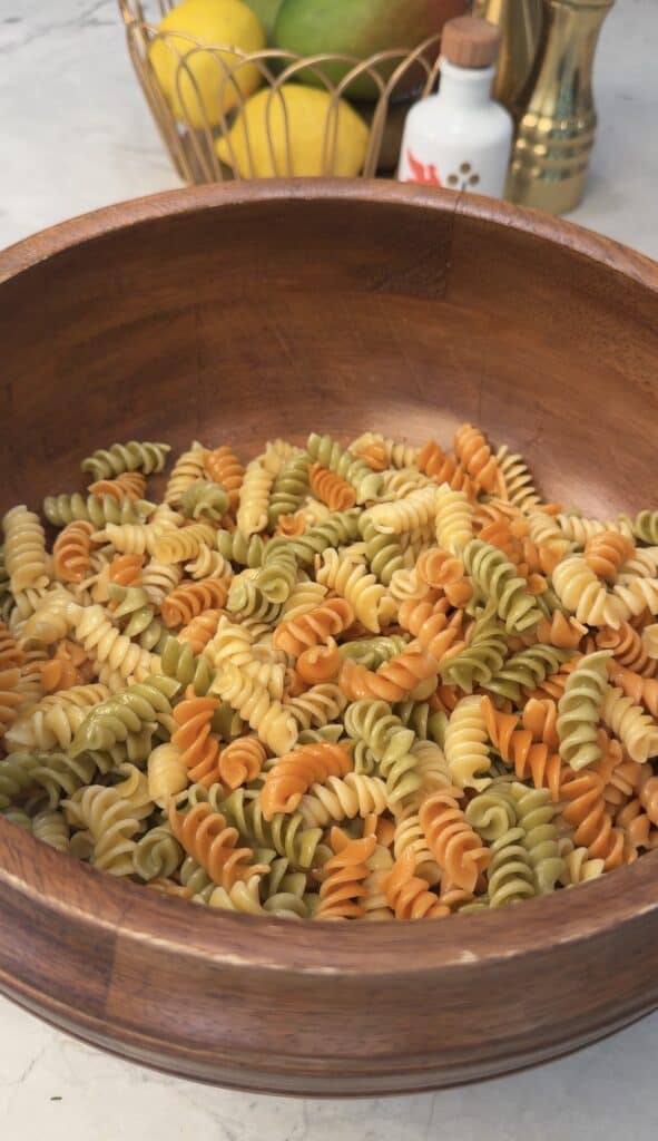 A wooden bowl filled with uncooked tri-color rotini pasta sits on a countertop, ready to be transformed into a high protein deviled egg pasta salad, with a fruit basket and seasonings visible in the blurred background.