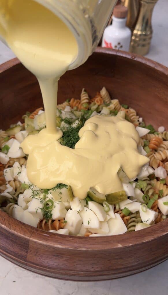 A creamy yellow dressing is being poured over a wooden bowl filled with tri-color pasta, chopped eggs, pickles, green onions, and fresh dill for a high protein deviled egg pasta salad.