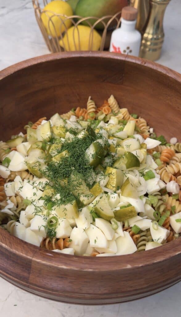 A wooden bowl filled with high protein deviled egg pasta salad featuring tri-color rotini pasta, chopped hard-boiled eggs, diced pickles, green onions, and fresh dill, set on a light surface.