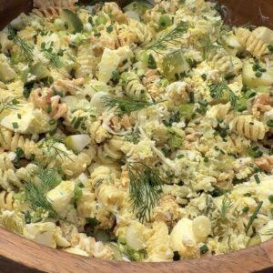 A wooden bowl filled with creamy, high protein deviled egg pasta salad, featuring rotini, sliced hard-boiled eggs, chives, dill, and green onions, set on a light surface.