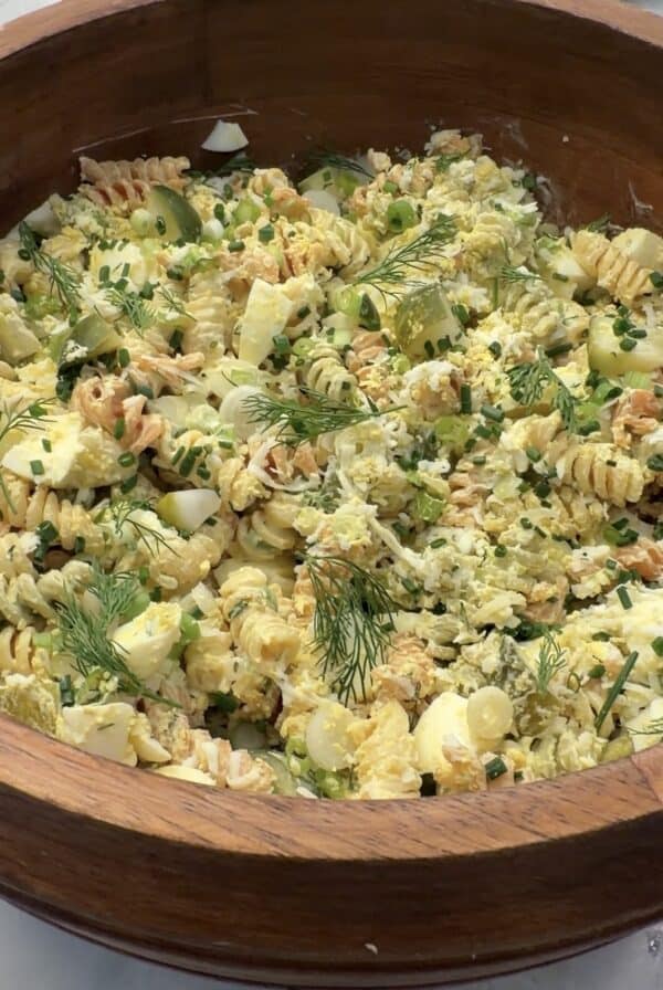 A wooden bowl filled with creamy, high protein deviled egg pasta salad, featuring rotini, sliced hard-boiled eggs, chives, dill, and green onions, set on a light surface.