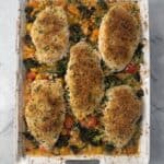 A Lazy One-Pan Chicken dinner dish with four breaded chicken breasts on top of mixed vegetables, including broccoli and cherry tomatoes, on a marble countertop.