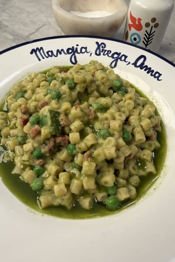 A plate of ditalini pasta with peas, zucchini, and a vibrant green sauce sits on a white dish that reads Mangia, Pregha, Ama. Reminiscent of classic Peas & Pancetta Pasta—though here served in a lighter vegetarian style—a salt cellar and a decorative bottle can be seen in the background.