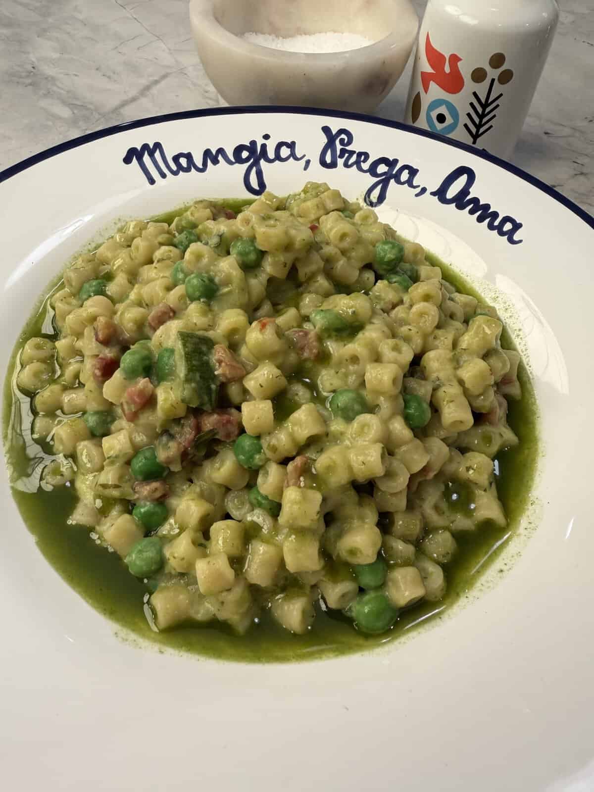 A plate of ditalini pasta with peas, zucchini, and a vibrant green sauce sits on a white dish that reads Mangia, Pregha, Ama. Reminiscent of classic Peas & Pancetta Pasta—though here served in a lighter vegetarian style—a salt cellar and a decorative bottle can be seen in the background.