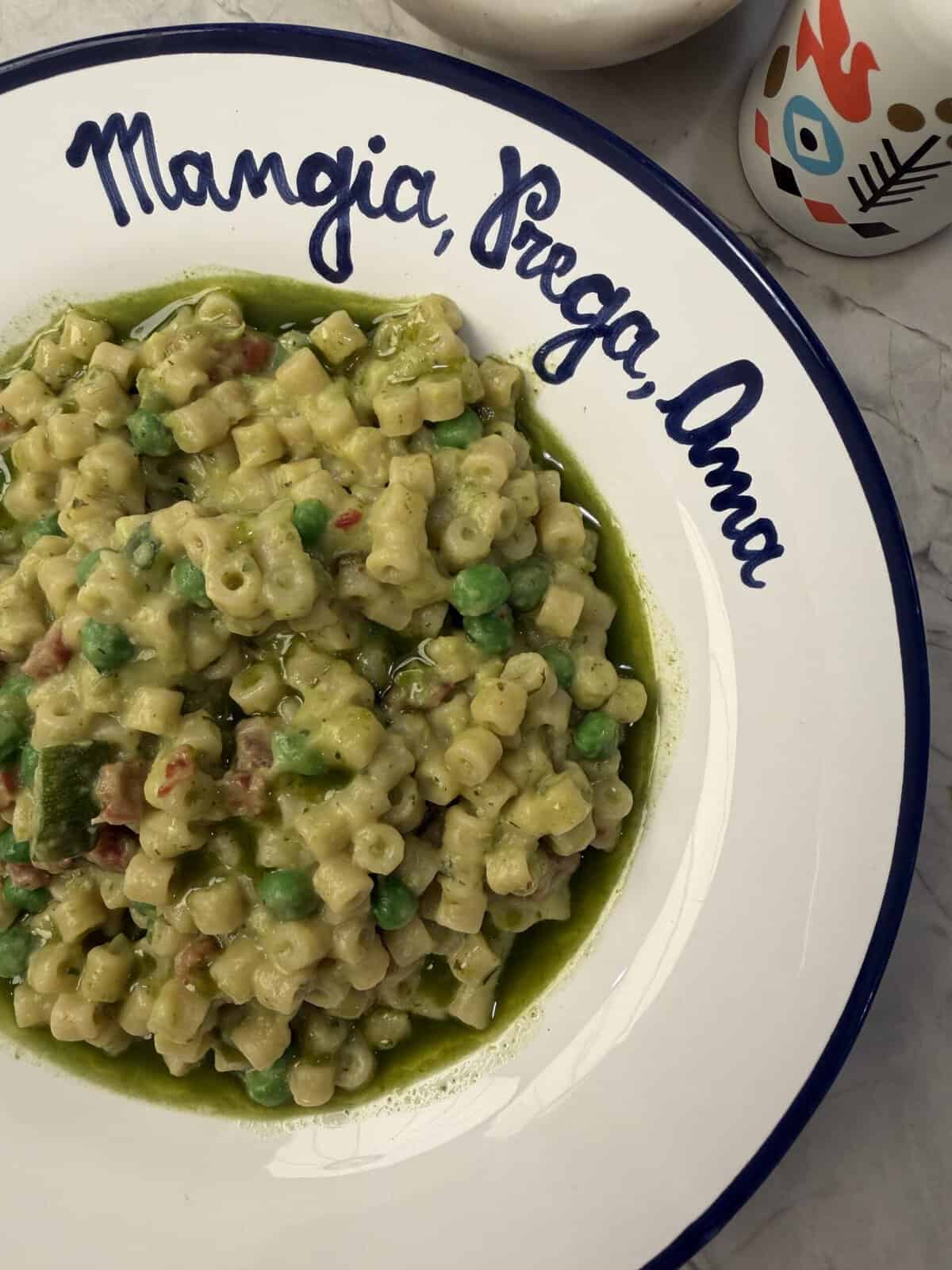 A white plate with blue writing holds creamy Peas & Pancetta Pasta, garnished with vibrant peas. The plate reads “Mangia, Prega, Ama.” Part of a white napkin and a decorative cup are visible on a marble surface.