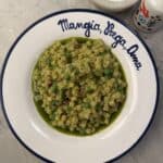 A bowl of Peas & Pancetta Pasta with green peas and herbs sits on a white plate that reads Mangia, Beve, Ama. A salt bowl and a decorative bottle are nearby on a marble surface.