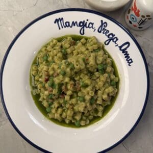 A bowl of Peas & Pancetta Pasta with green peas and herbs sits on a white plate that reads Mangia, Beve, Ama. A salt bowl and a decorative bottle are nearby on a marble surface.