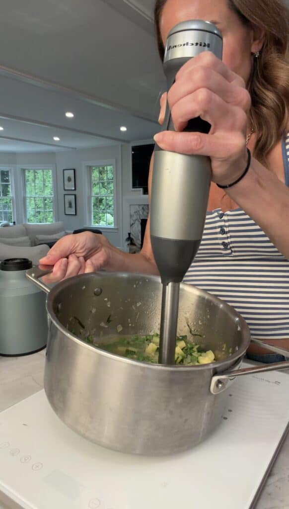 A woman in a striped tank top uses an immersion blender to mix ingredients for Peas & Pancetta Pasta in a large pot on a kitchen counter.