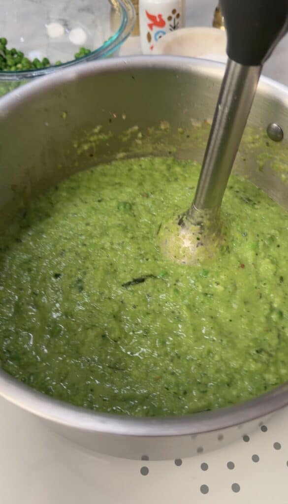 A metal pot filled with green vegetable soup, reminiscent of the vibrant flavors found in Peas & Pancetta Pasta, is being blended with an immersion blender.