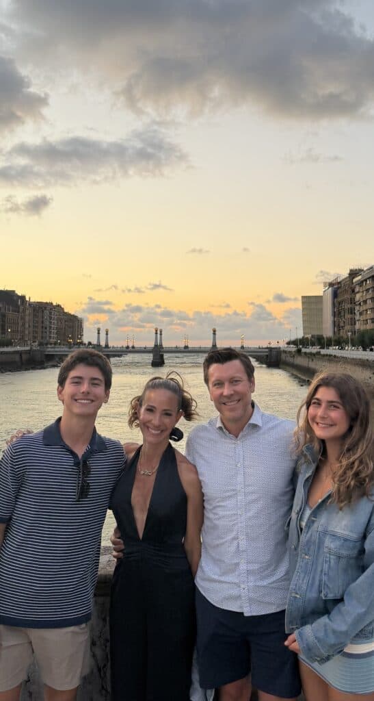 Four people smile together by a river at sunset, with buildings lining both sides and a bridge in the background under a cloudy sky—capturing just one of the memorable moments you might experience while exploring What To Do In San Sebastian.