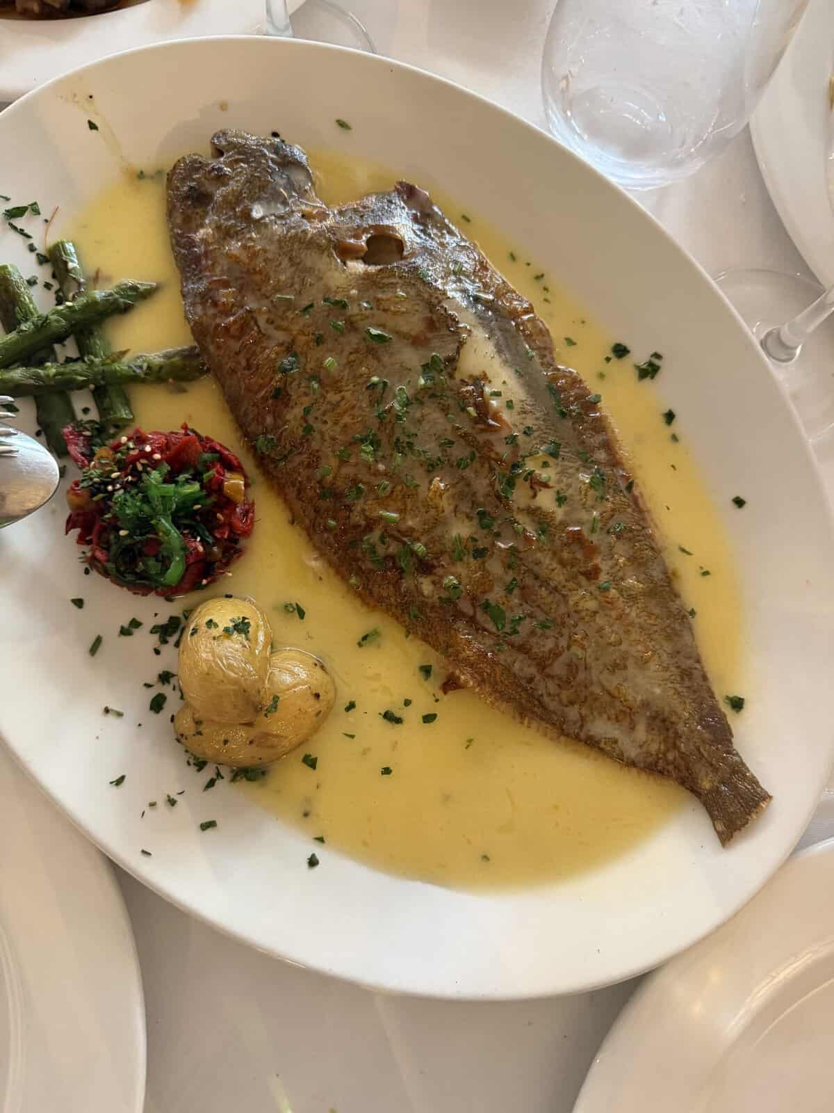 A whole cooked fish in yellow sauce garnished with herbs, served with two potatoes, green beans, and a small red vegetable mix on a white plate, with glasses visible in the background—a dish that perfectly captures the culinary spirit of What To Do In San Sebastian.