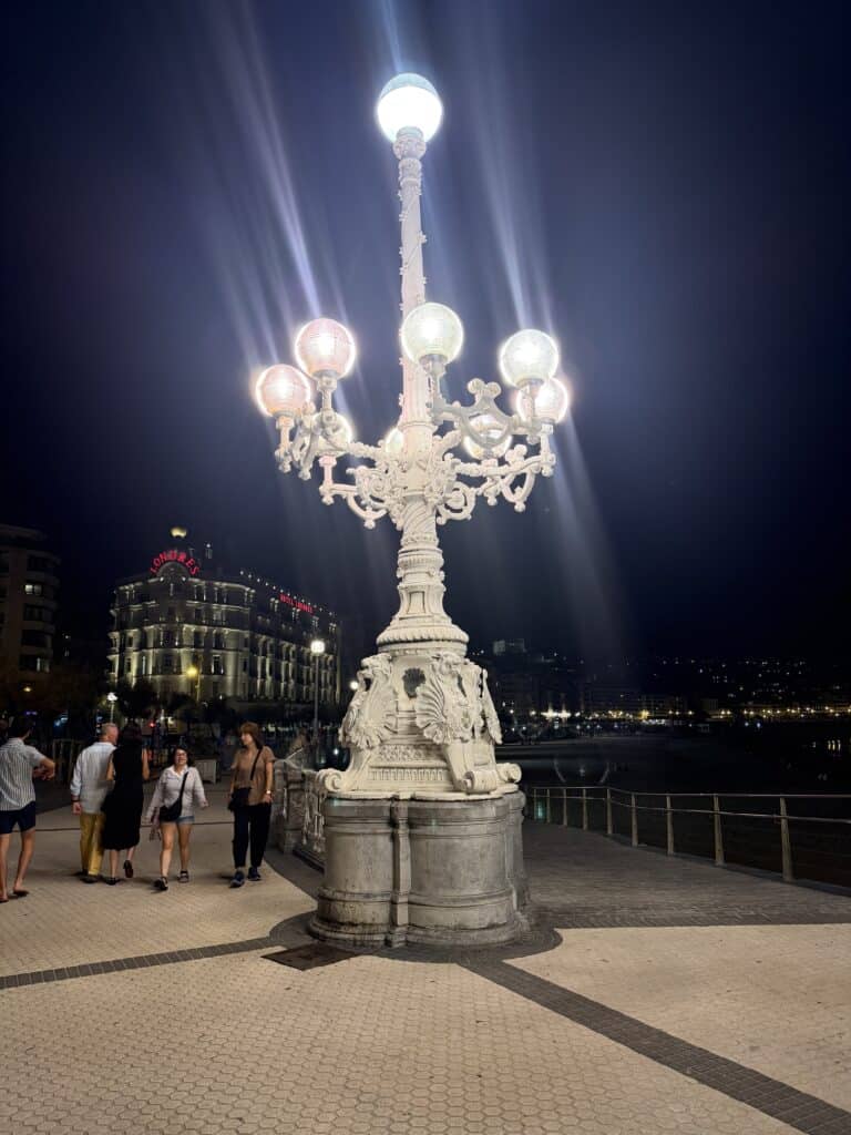 A large ornate streetlamp with glowing bulbs stands on a riverside promenade at night—a quintessential scene for anyone exploring What To Do In San Sebastian. People walk nearby, soaking in the atmosphere, while a grand illuminated building is visible in the background.