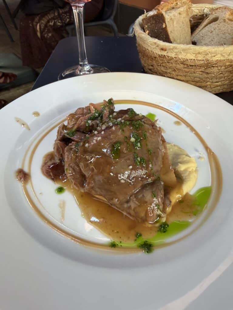 A plate with braised meat in brown sauce, garnished with herbs and served over mashed potatoes—a true comfort dish you might discover when exploring What To Do In San Sebastian. In the background, a basket of sliced bread and a glass of wine complete this inviting scene.