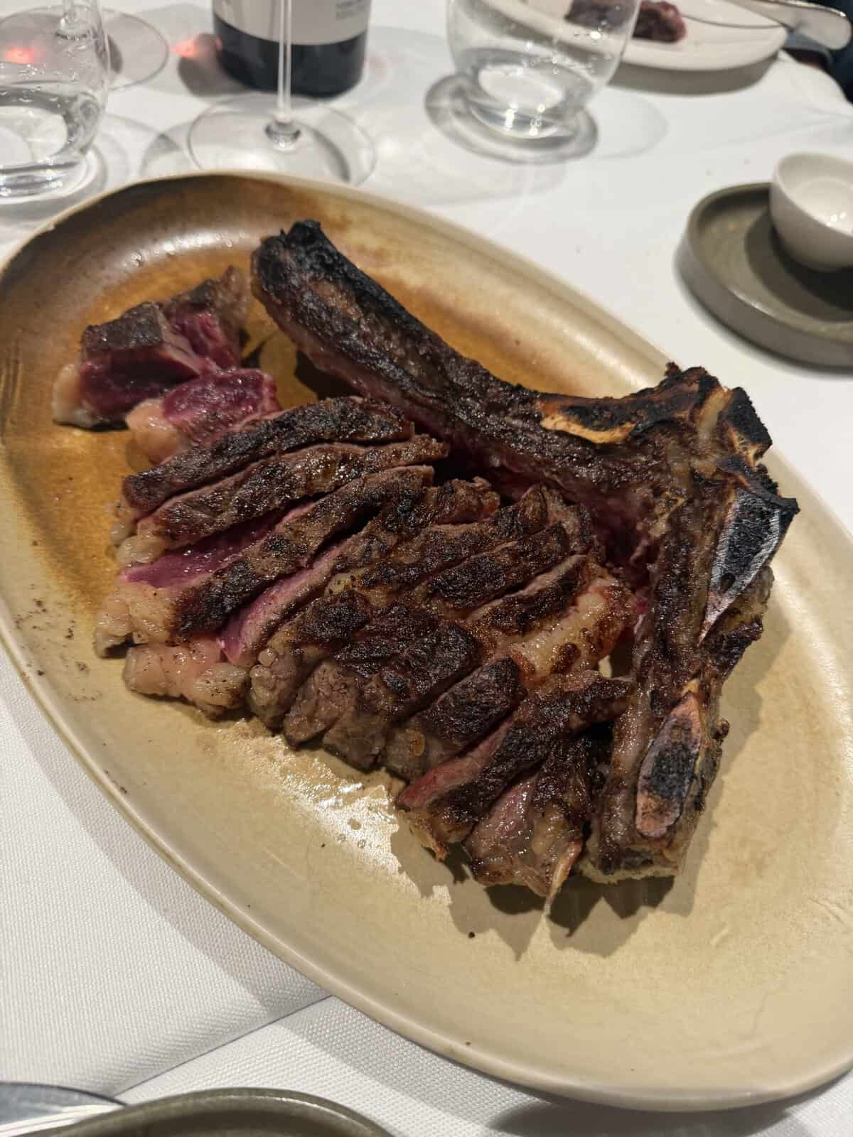 A sliced, medium-rare tomahawk steak is served on an oval plate, showing a seared crust and juicy pink interior. Empty glasses and small plates surround it on a white tablecloth—a scene reminiscent of the lively dining experiences found when exploring What To Do In San Sebastian.