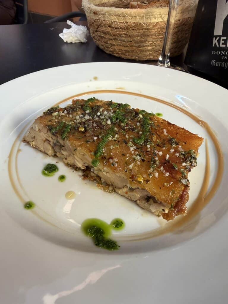 A slice of roasted pork with crispy skin, garnished with herbs and sauce, is served on a white plate with green oil drizzles—a mouthwatering example of What To Do In San Sebastian for food lovers. A basket and a napkin can be seen in the background.