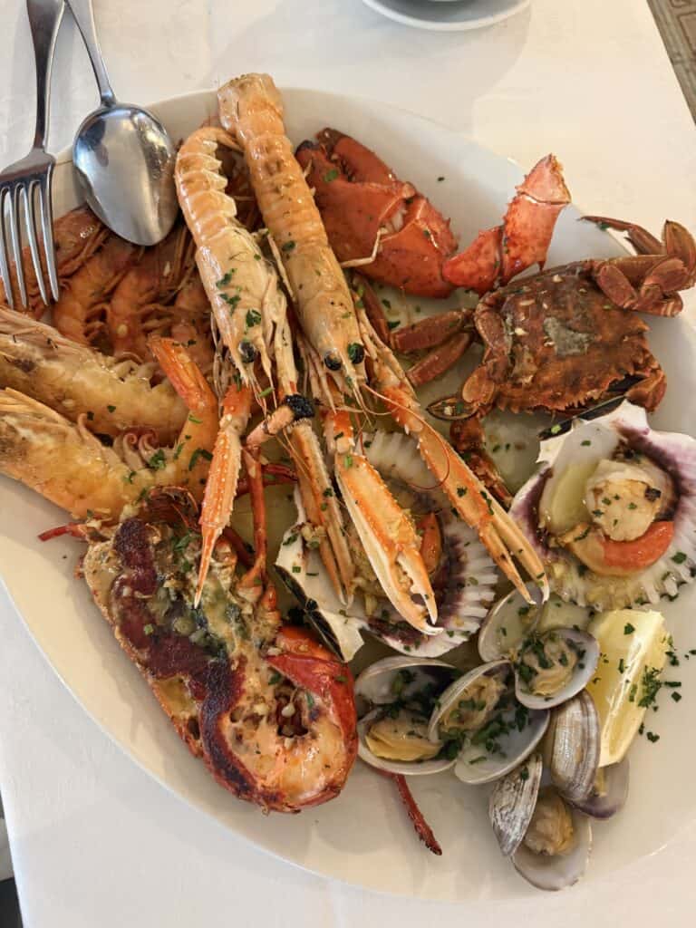 A platter of assorted seafood including shrimp, lobster, crab, clams, scallops, and prawns, garnished with herbs, served on a white plate with utensils beside it—an experience that highlights exactly What To Do In San Sebastian for food lovers.