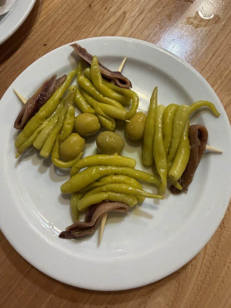 White plate with skewers of green peppers, green olives, and anchovies on a wooden table. The food is arranged in a circular pattern with three skewers visible—a classic pintxo you might find while exploring What To Do In San Sebastian.