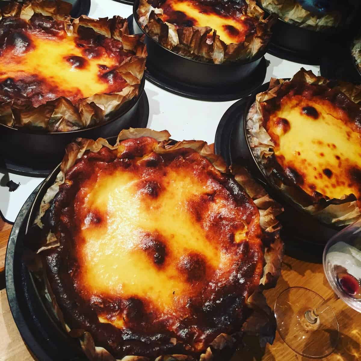 Several Basque burnt cheesecakes in round pans with browned, caramelized tops sit on a counter—an iconic treat and a must-try for anyone wondering What To Do In San Sebastian. A wine glass with red wine is visible nearby. The cheesecakes are on baking paper, slightly charred at the edges.