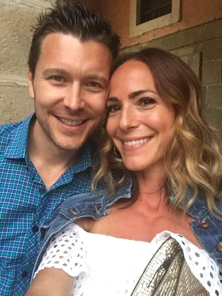 A smiling man in a blue checkered shirt and a woman in a white off-the-shoulder top pose for a selfie together in front of a stone wall, capturing the joy of discovering What To Do In San Sebastian.