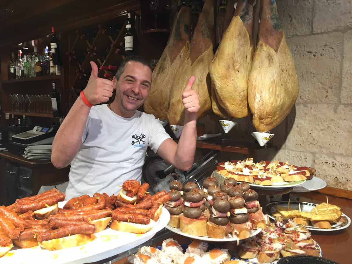 A smiling man gives two thumbs up behind a bar showcasing various tapas, including sausages, stuffed mushrooms, and other appetizers, with large hanging cured hams in the background—a perfect glimpse into What To Do In San Sebastian for food lovers.