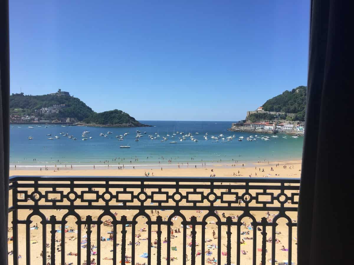 View from a balcony overlooking a sandy beach with people, calm blue sea, and numerous boats—a perfect introduction to What To Do In San Sebastian. Hills with greenery and buildings are visible on both sides under a clear blue sky, inviting you to explore the city’s stunning natural beauty and vibrant coastal life.