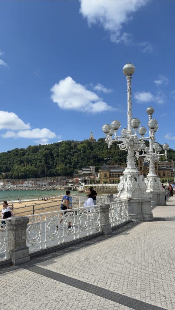 Ornate white railings and lamp posts line a lively seaside promenade in San Sebastian, with people strolling along and soaking in the atmosphere. A sandy beach stretches alongside the walkway beneath a blue sky dotted with clouds, while a lush green hill crowned by a statue provides a stunning backdrop—perfect inspiration for anyone searching for what to do in San Sebastian.