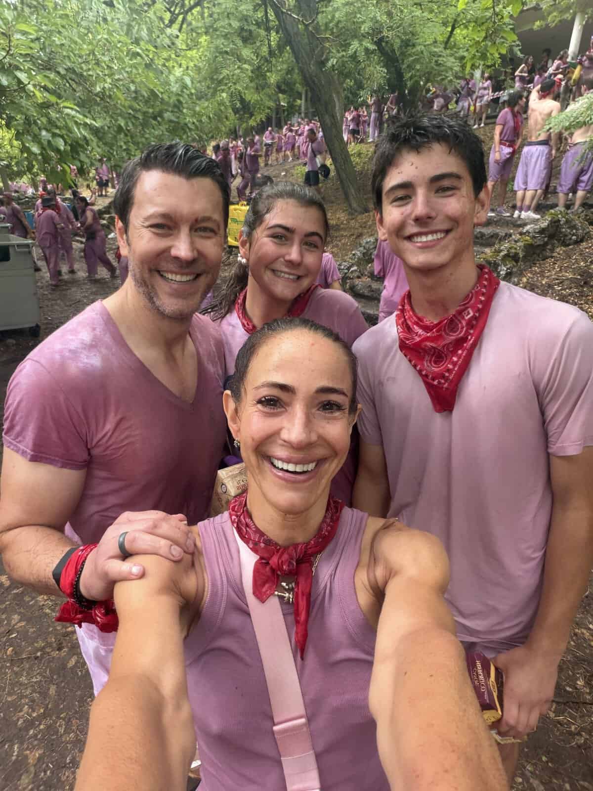 Four people in matching purple shirts and red bandanas smile for a selfie outdoors, capturing the festive spirit of a local event. Many others in similar outfits gather in the background among trees and greenery, offering a glimpse into what to do in San Sebastian for those eager to experience vibrant community traditions.