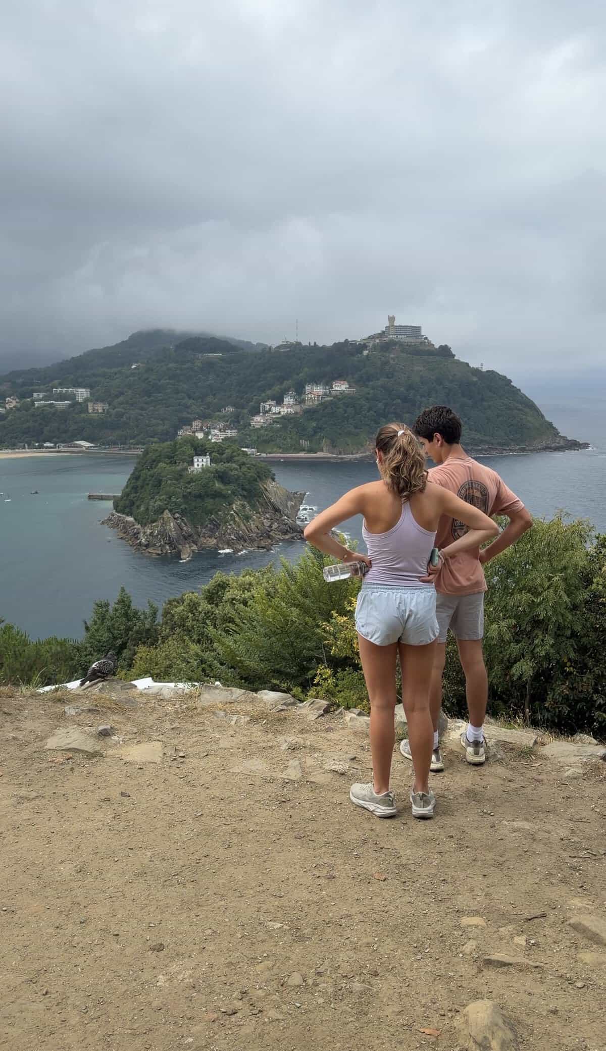 Two people stand on a cliffside path overlooking the ocean, with lush green islands and cloudy skies in the distance—a peaceful and slightly overcast scene that captures the serene beauty of exploring coastal trails, one of the highlights of What To Do In San Sebastian.