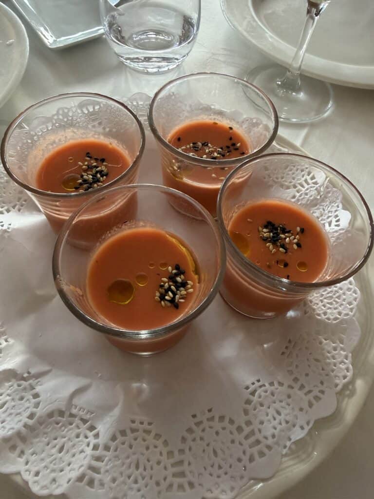 Four clear glasses filled with orange gazpacho soup, garnished with seeds and olive oil, placed on a white doily-covered plate on a table set for a meal—just one of the delicious culinary experiences you might discover while exploring What To Do In San Sebastian.