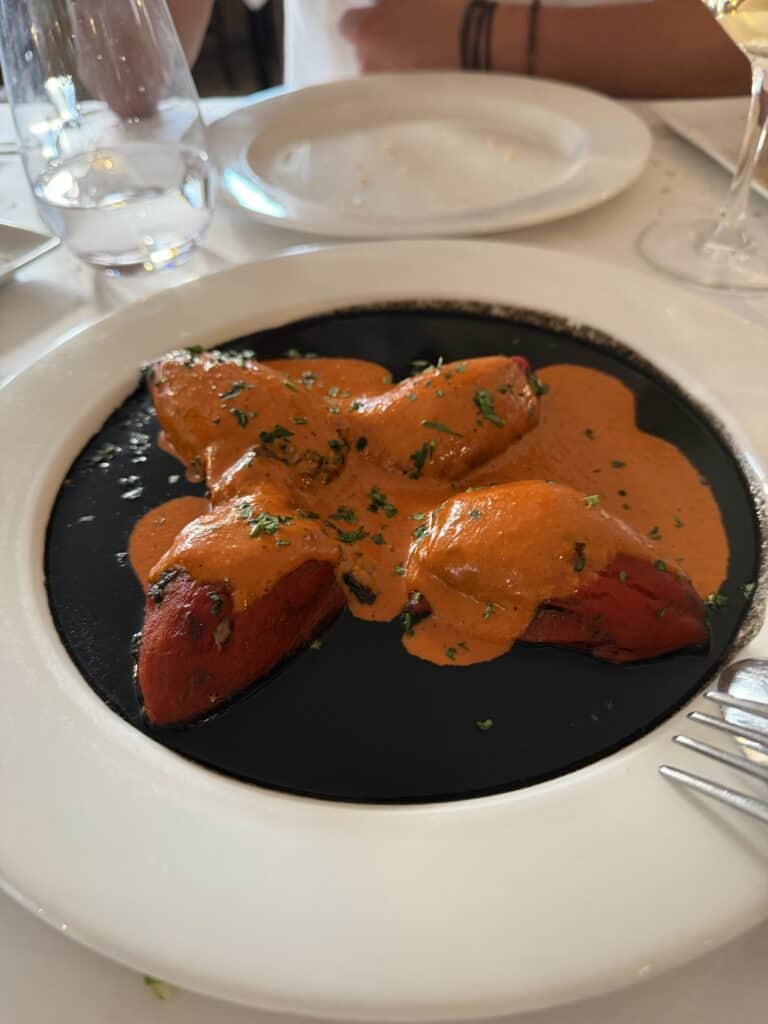 Three stuffed red peppers topped with orange sauce are served on a black plate, with a fork visible—an inviting dish reminiscent of the vibrant culinary experiences you’ll find while exploring What To Do In San Sebastian. A glass of water and white plates are in the background, completing this appetizing scene.