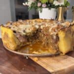 A savory Steak & Potato Pie sits on a wooden board with a slice removed, revealing its cheesy, meaty filling. A floral arrangement and brass candlestick are in the blurred background.