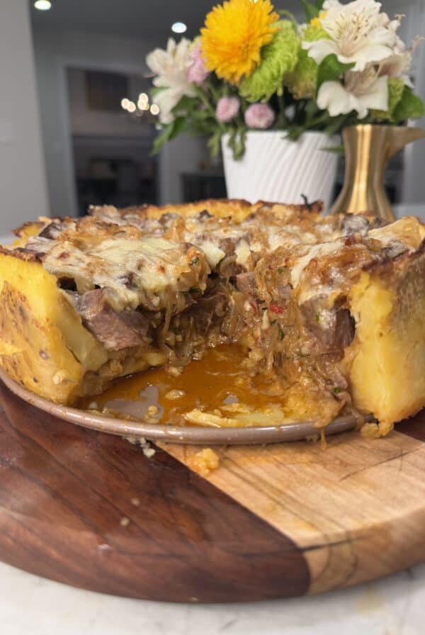 A savory Steak & Potato Pie sits on a wooden board with a slice removed, revealing its cheesy, meaty filling. A floral arrangement and brass candlestick are in the blurred background.