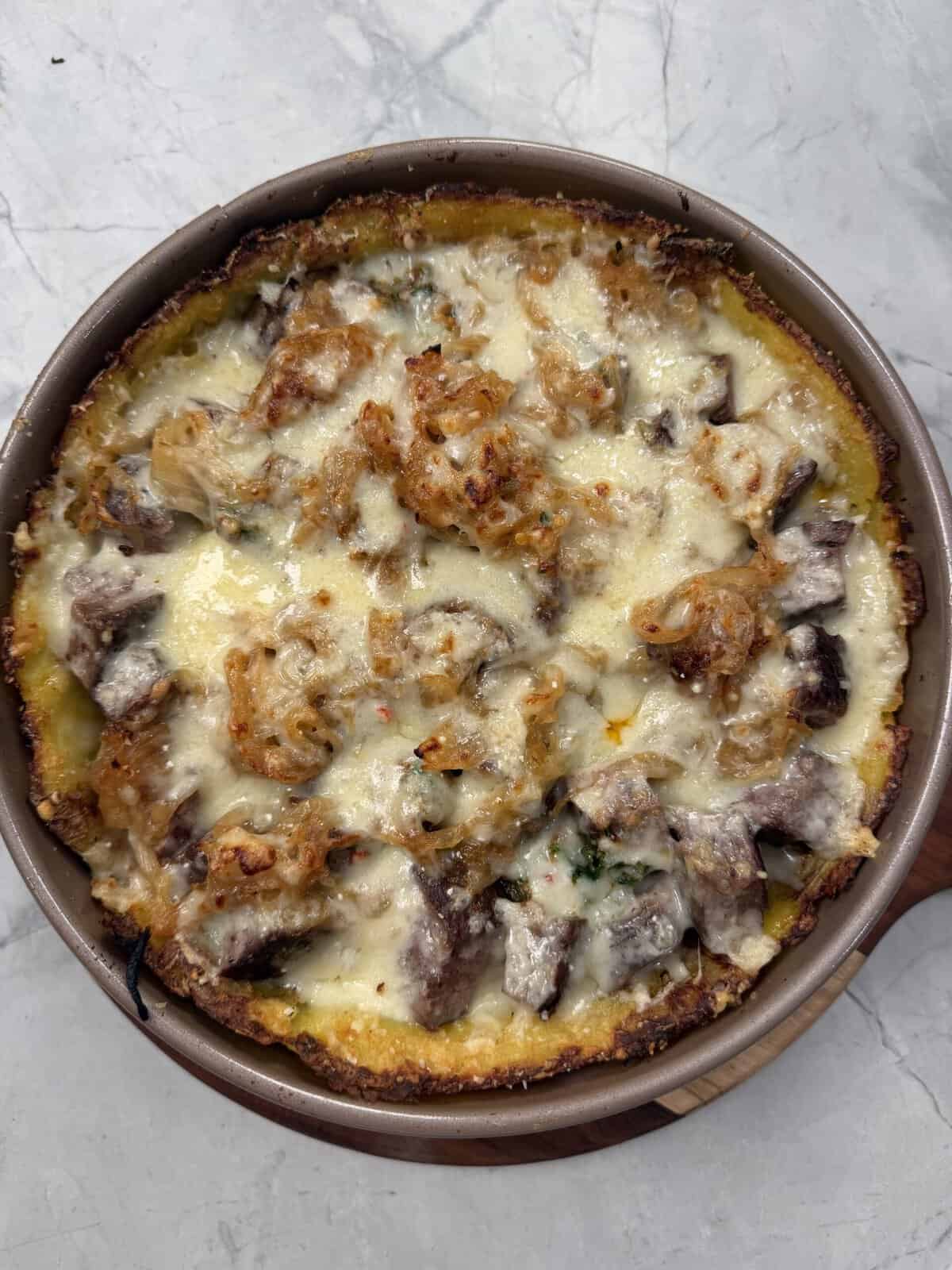 A round baked dish filled with melted cheese, browned beef or sausage pieces, and herbs rests on a marble surface, reminiscent of a classic Steak & Potato Pie.