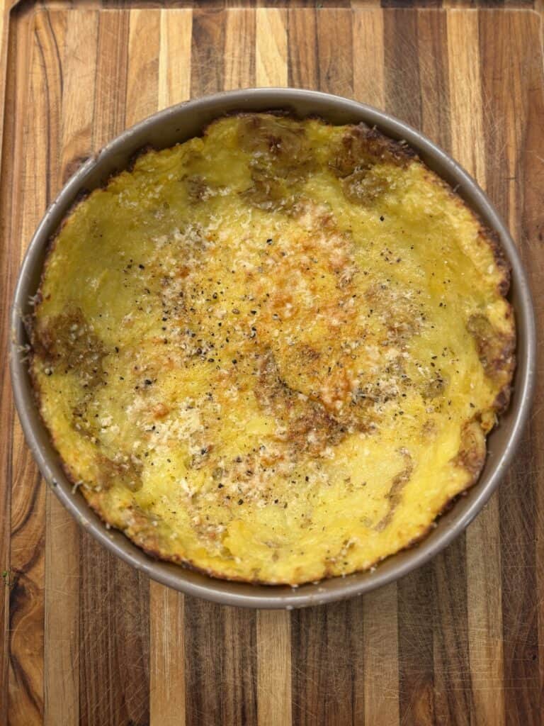 A round baking dish filled with a golden, crispy mashed potato crust, lightly sprinkled with black pepper and grated cheese, sits on a wooden cutting board—perfect for serving a classic Steak & Potato Pie.