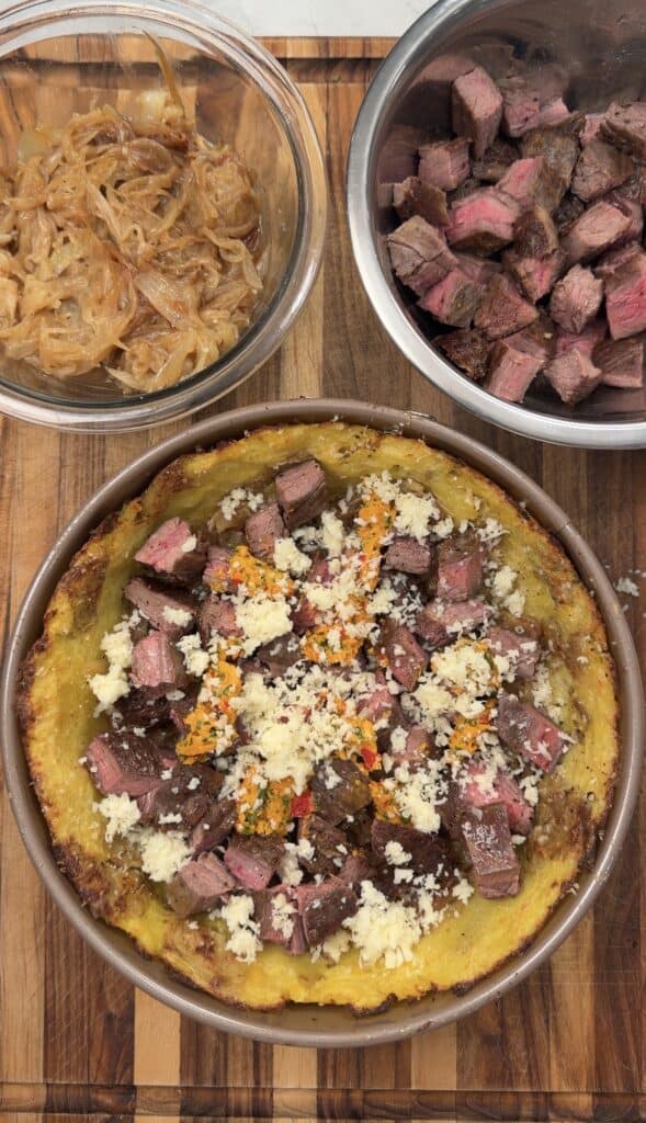 A hearty Steak & Potato Pie sits on a wooden board, topped with diced steak, grated cheese, and a rich sauce. Nearby are bowls of caramelized onions and extra diced steak, ready to pile onto this savory classic.