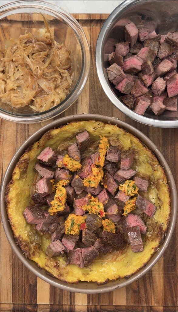 A baked Steak & Potato Pie dish topped with steak cubes and herb butter, surrounded by bowls of caramelized onions and diced steak, all on a wooden cutting board.